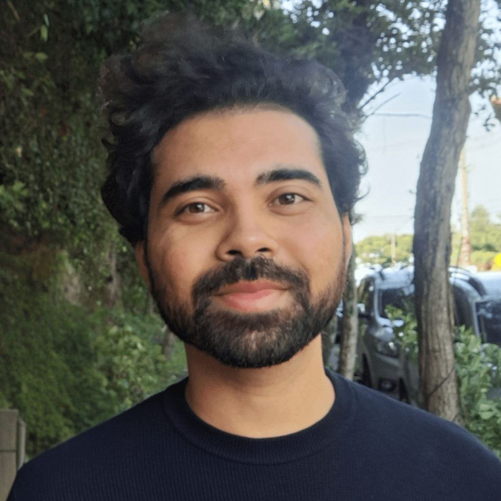A smiling man with curly hair and a beard, wearing a dark sweater, standing outdoors with greenery and parked cars in the background.