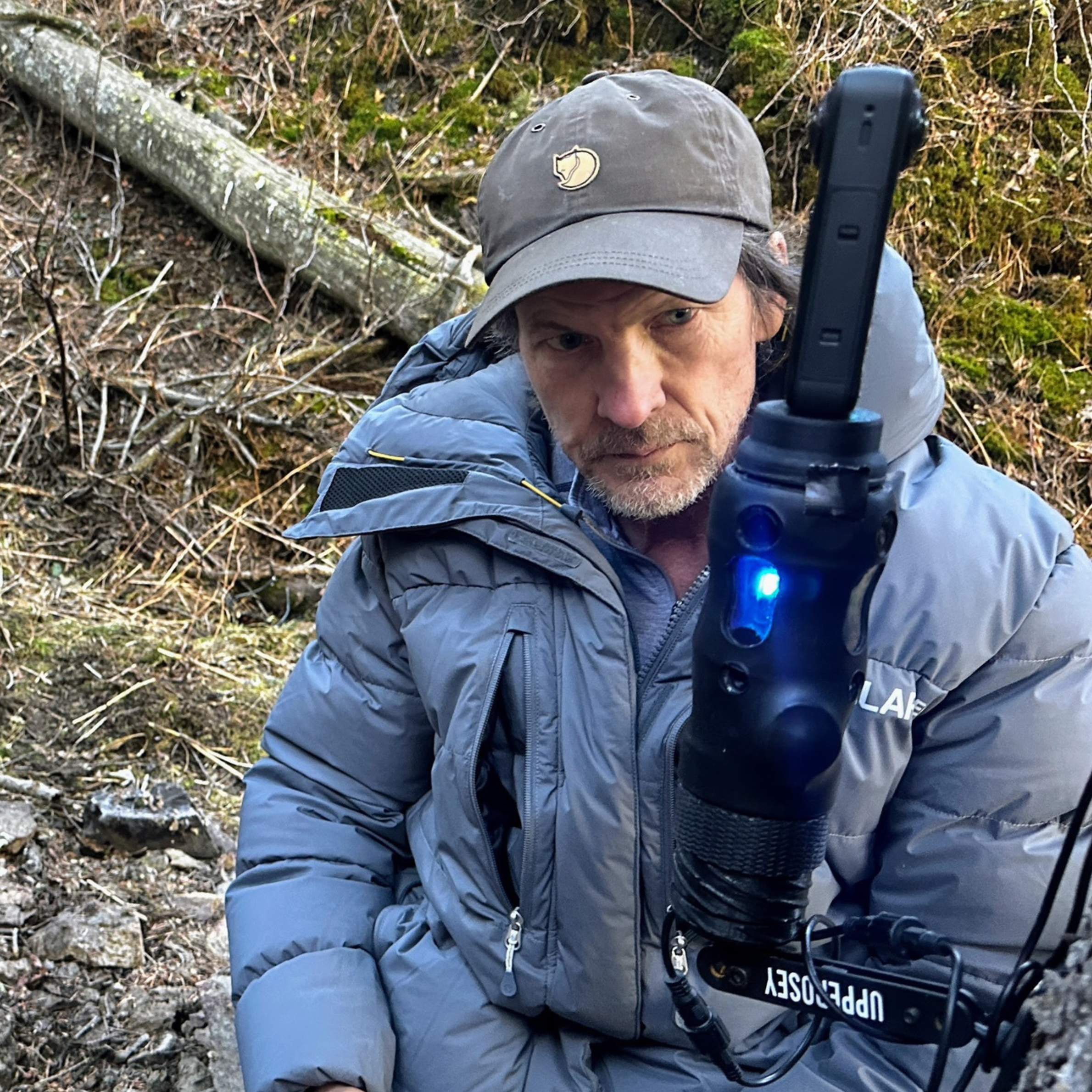 A man wearing a gray puffer jacket and a cap is sitting outdoors, focused on a device with a blue light. The background features natural elements like moss, tree branches, and rocks.